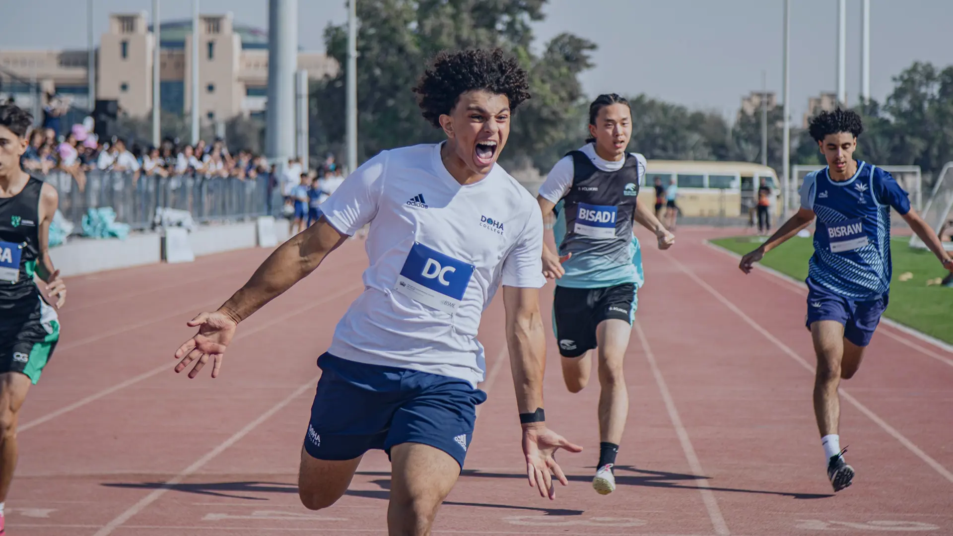 A Doha College pupil sprints across the finish line during an athletics race, celebrating with visible excitement as competitors follow behind on the track.
