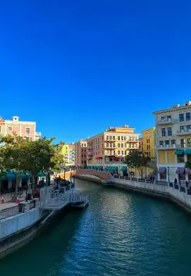 A canal lined with pastel-coloured buildings and shopfronts at Qanat Quartier in The Pearl, Doha, with clear blue skies overhead.